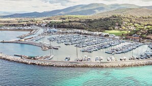 A marina with numerous boats docked, surrounded by a rocky breakwater and a backdrop of hills and greenery.