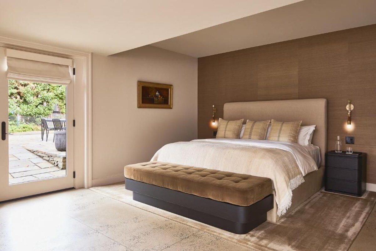 A bedroom with a large bed, a bedside table, a bench, and a door leading to an outdoor area.
