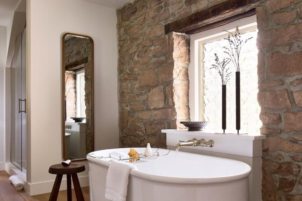 A bathroom with a stone wall, a large mirror, a bathtub, and a window with a vase of flowers.