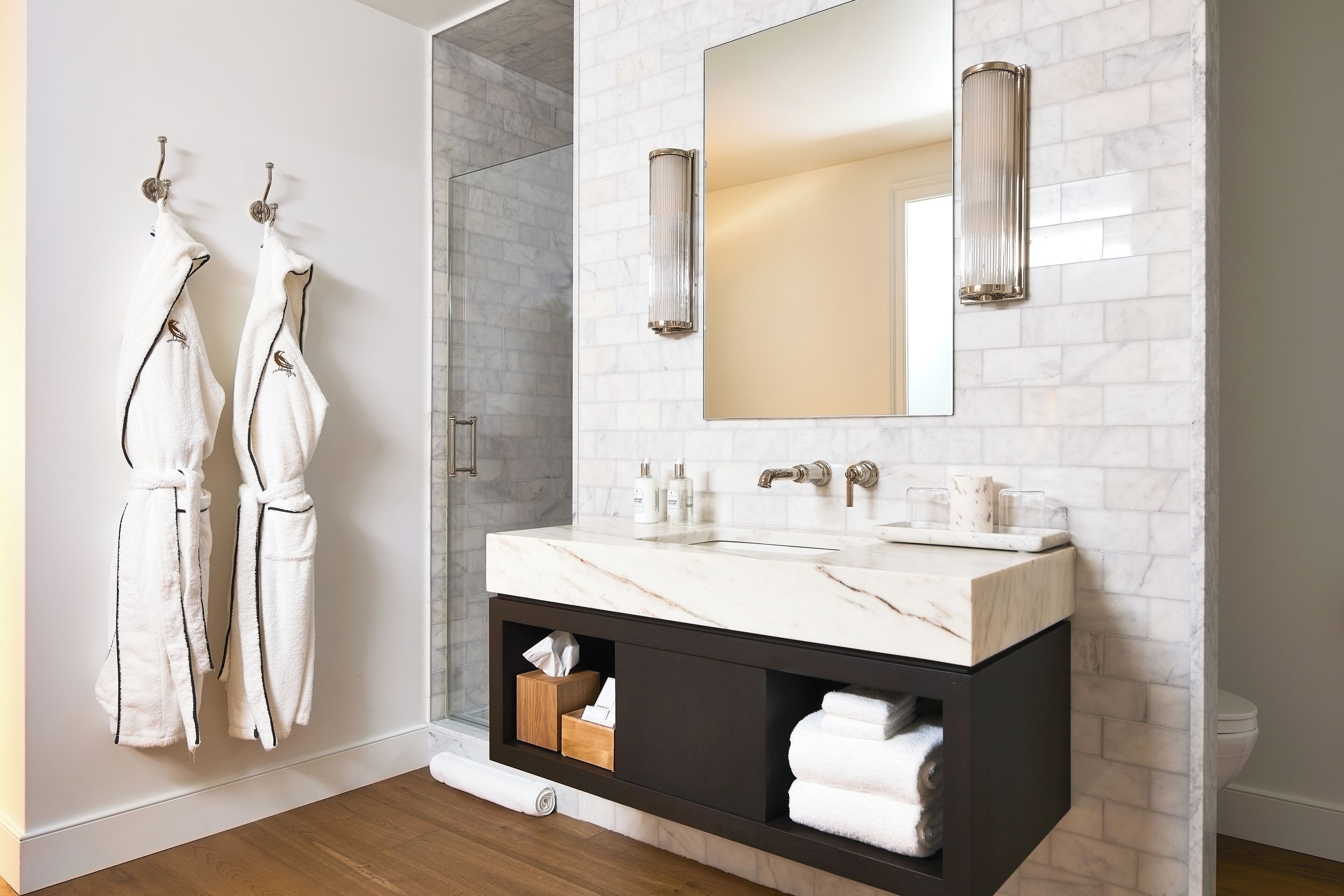 Un baño moderno con un lavamanos de mármol, dos luces empotradas en la pared y un espejo. Hay dos albornoces colgados en ganchos y un perchero con toallas.
