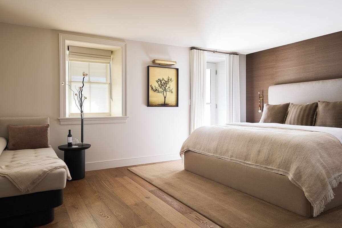 Ein Schlafzimmer mit einem großen Bett, einer kleinen Sitzecke, einem Fenster mit Jalousien, einem gerahmten Bild an der Wand und einem Nachttisch mit Flasche und Glas.