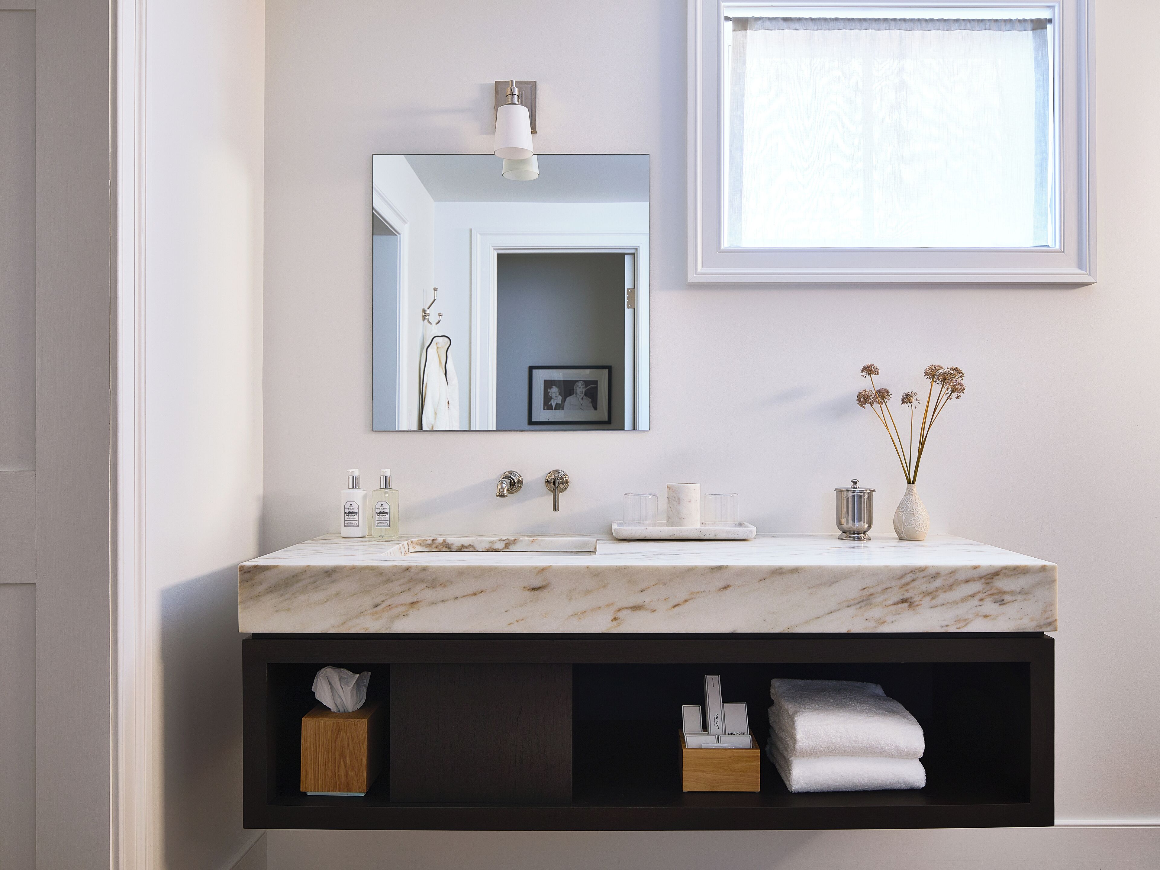 A bathroom with a marble sink, a mirror, a window with a white curtain, and a vase with dried flowers.