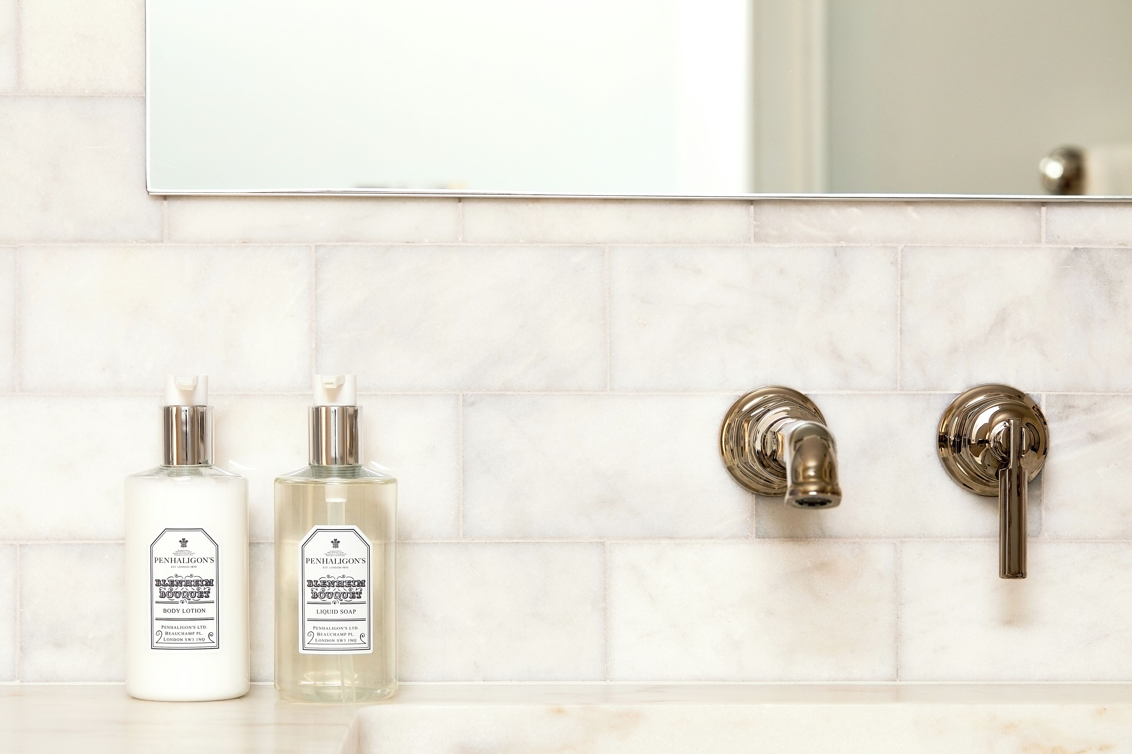 Dos botellas de loción corporal y dos grifos de cromo en un tocador de baño con un salpicadero de azulejos blancos.