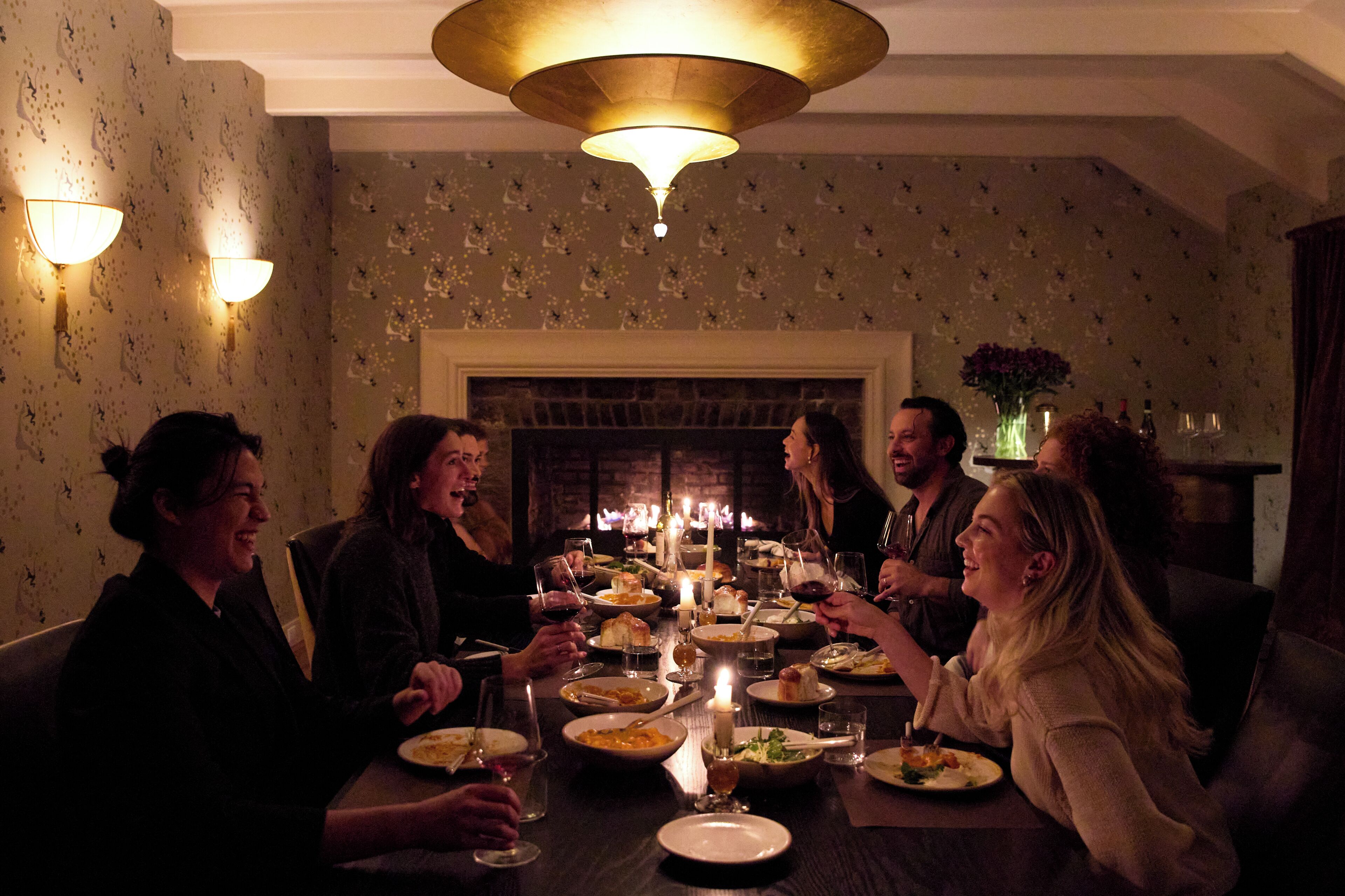 Un groupe de personnes est assis à une table, dînant à la lueur de bougies, entouré d’une cheminée et de luminaires muraux.