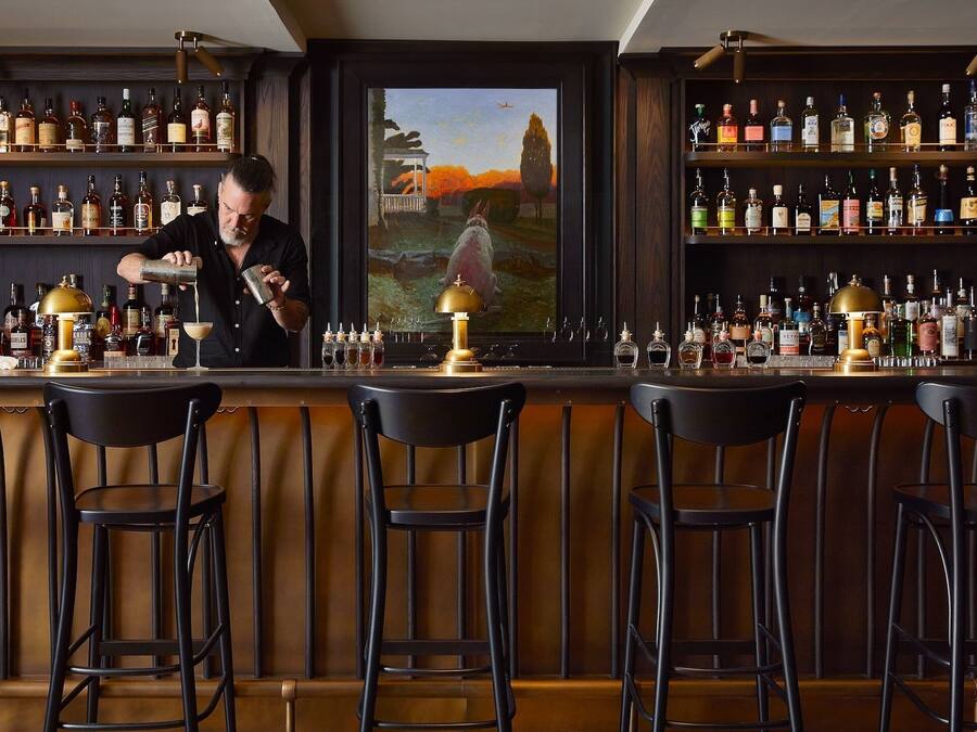 Um bar com um bartender preparando uma bebida, prateleiras cheias de garrafas de bebidas e uma pintura de uma paisagem.