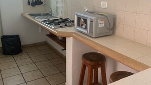 A kitchen with a gas stove, a microwave, and a sink. There are hanging utensils above the sink and a trash bag on the floor. - Bungalows Adelamar (Zihuatanejo)