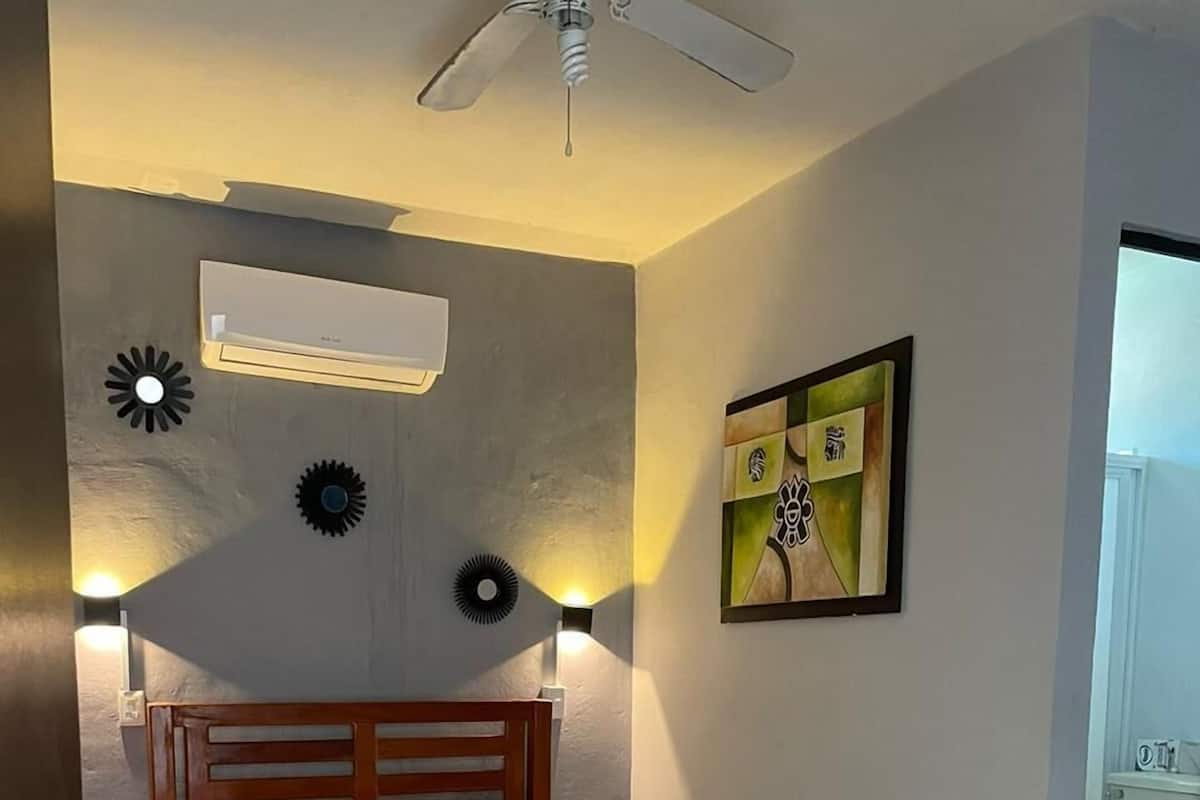 A hotel room with a bed, a ceiling fan, a wall-mounted air conditioner, and a painting.