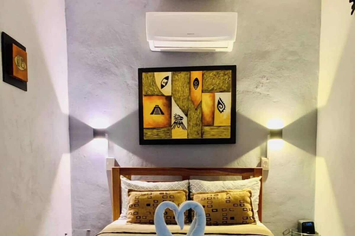 A hotel room with a bed, two wall-mounted lights, a painting, and an air conditioning unit.