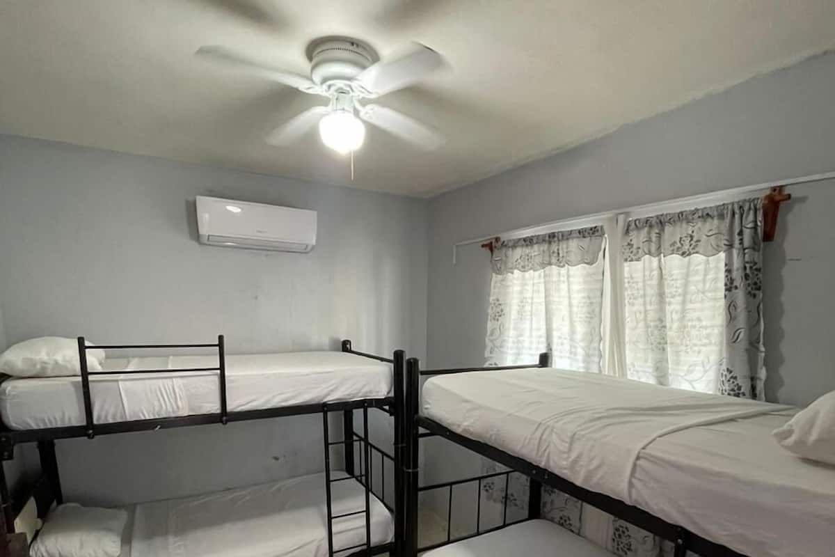 A dormitory room with bunk beds, an air conditioning unit, a ceiling fan, and a window with curtains.