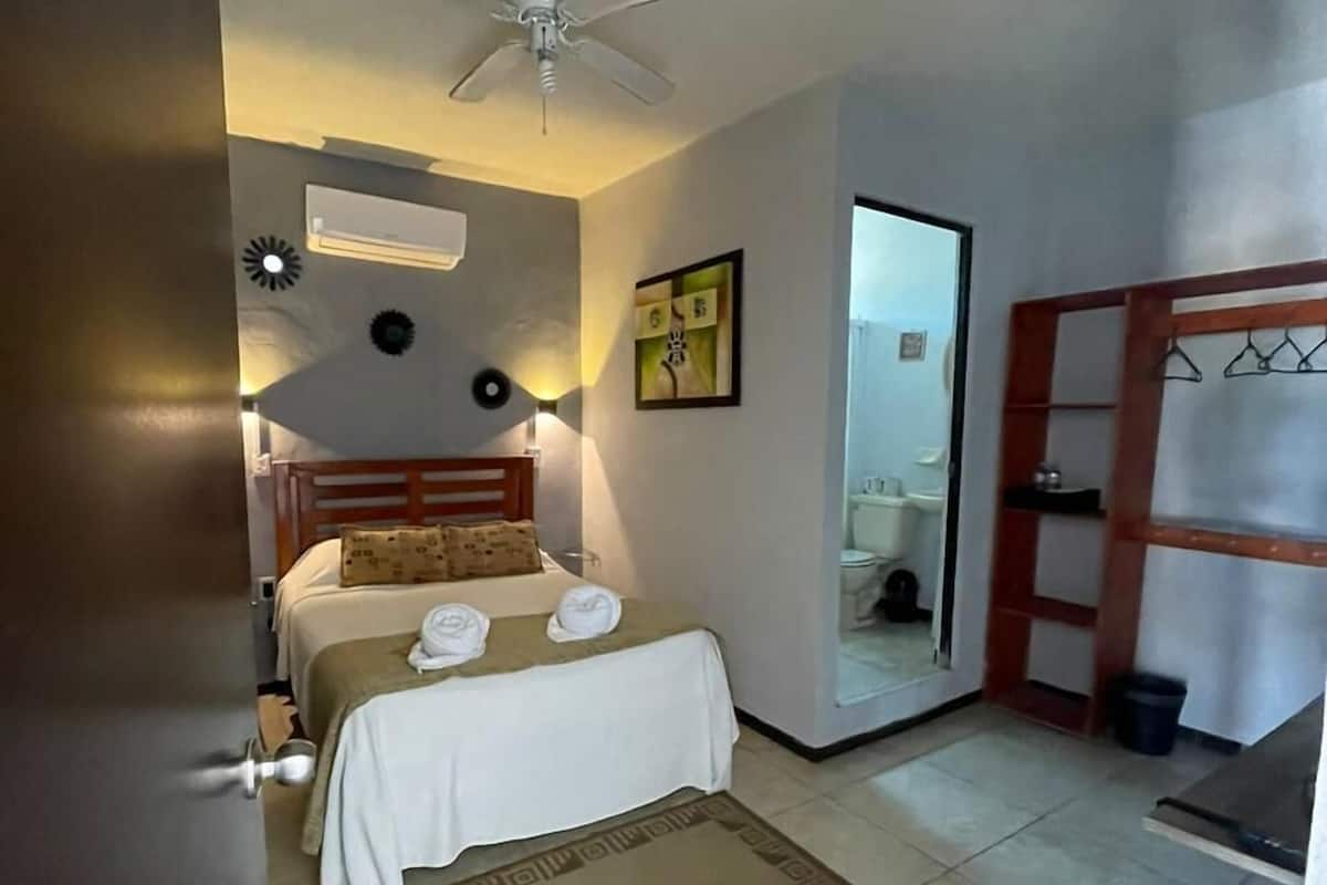 A hotel room with a bed, a ceiling fan, a bedside table, a nightstand, a mirror, a wardrobe, and a bathroom visible through an open door.