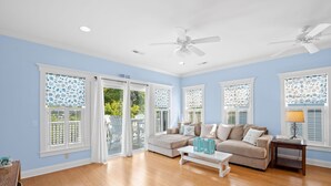 Ein geräumiges Wohnzimmer mit einem beigefarbenen Ecksofa, einem weißen Couchtisch und einem Beistelltisch mit Lampe. Zwei Deckenventilatoren, eine Glastür mit Schiebeelementen zu einer Außenpatio und ein Fenster mit gemusterten Jalousien sind ebenfalls vorhanden.