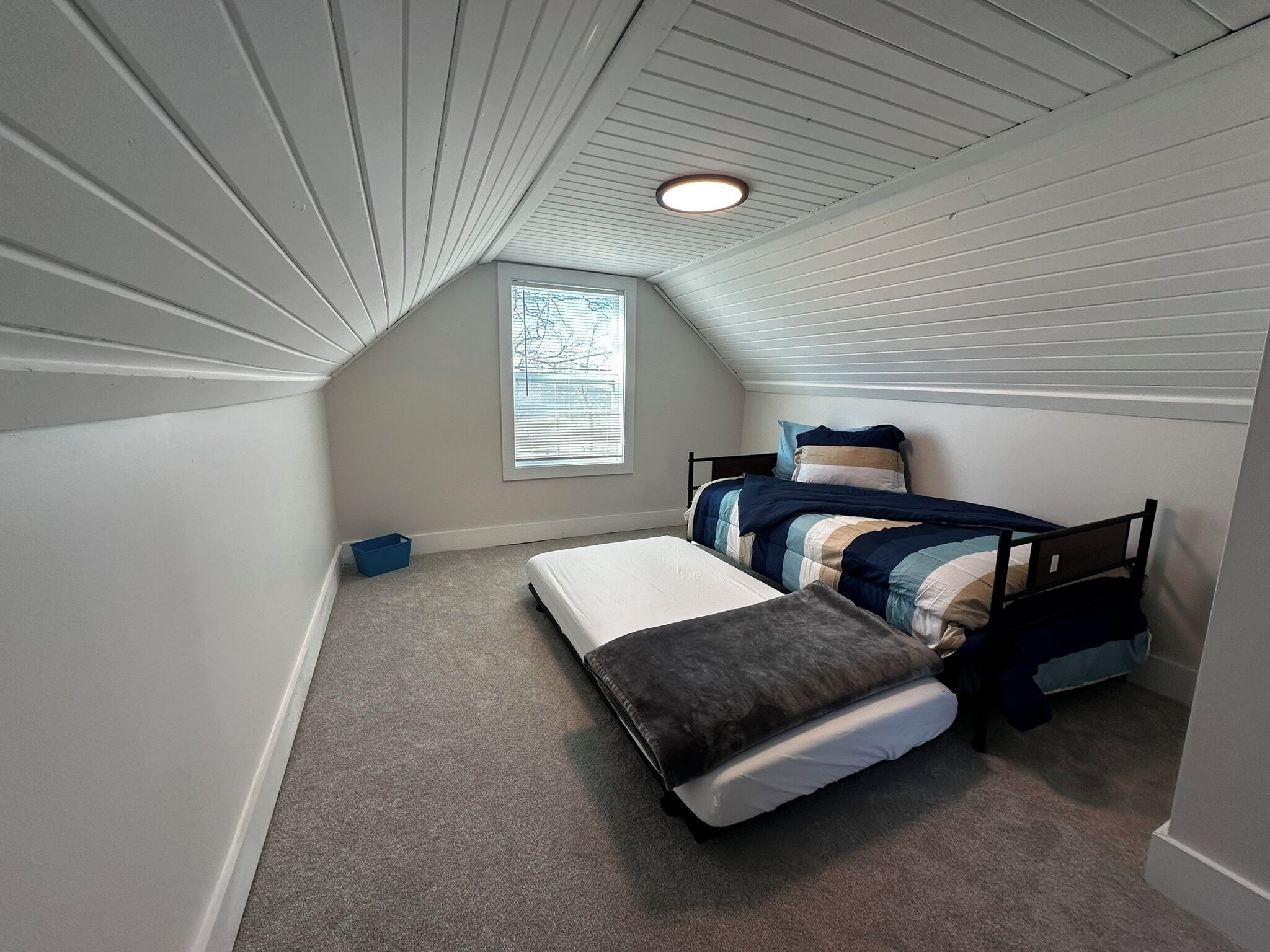 A small, sloped ceiling room with two beds, a window, and a blue storage bin.