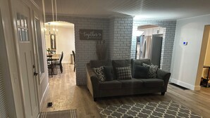 Un salon avec un canapé gris, un tapis à motifs et un mur en briques apparentes.
