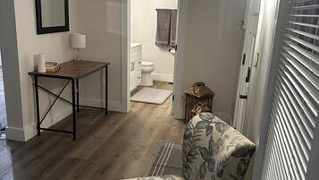 Un espacio habitable pequeño con suelo de madera, un sillón estampado con motivos florales, una mesita con una lámpara y una puerta que da a un baño.