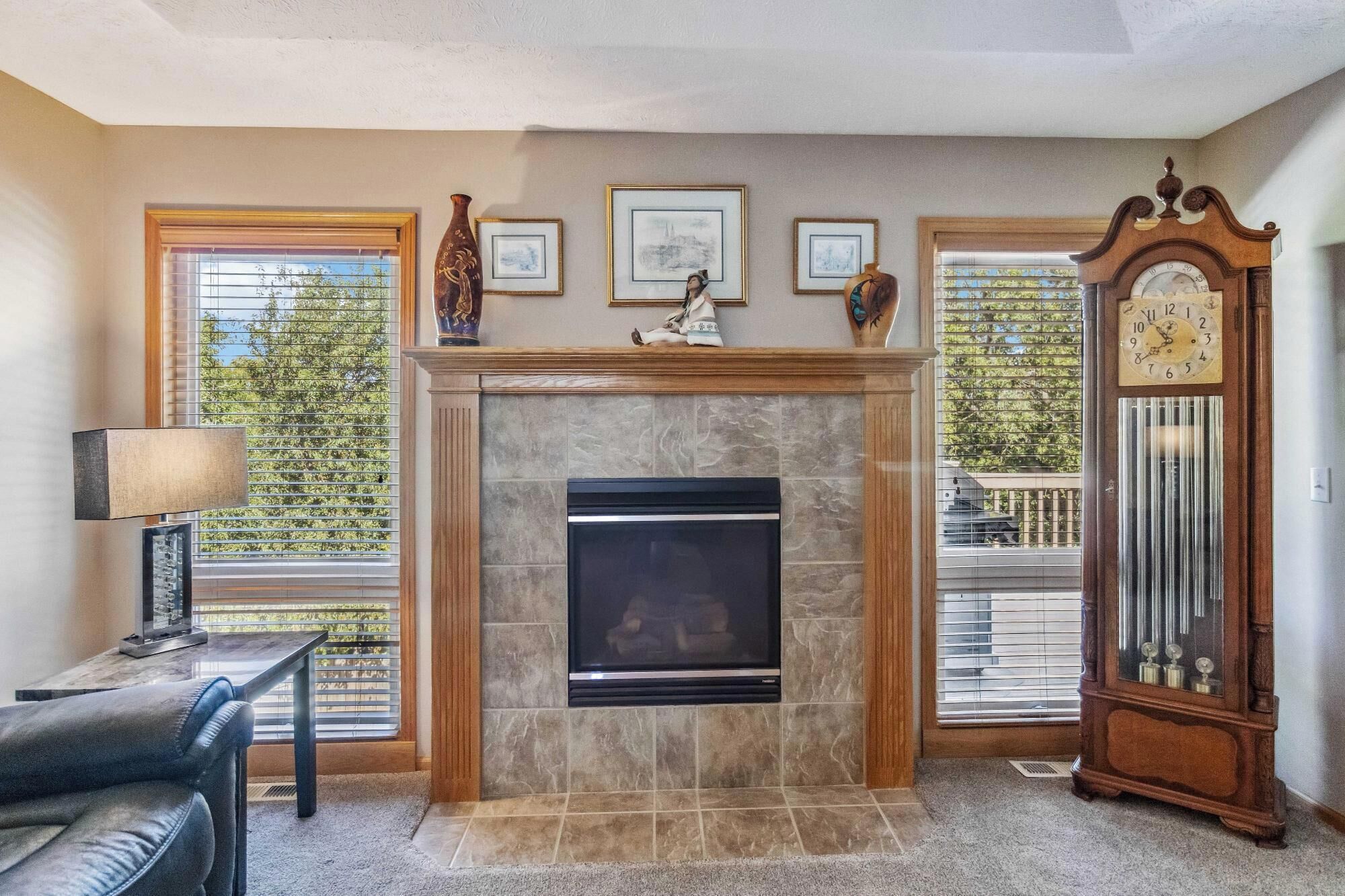 A living room with a fireplace, framed pictures, a clock, and a view of the outdoors.