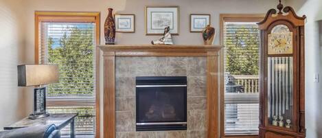 A living room with a fireplace, framed pictures, a clock, and a view of the outdoors.