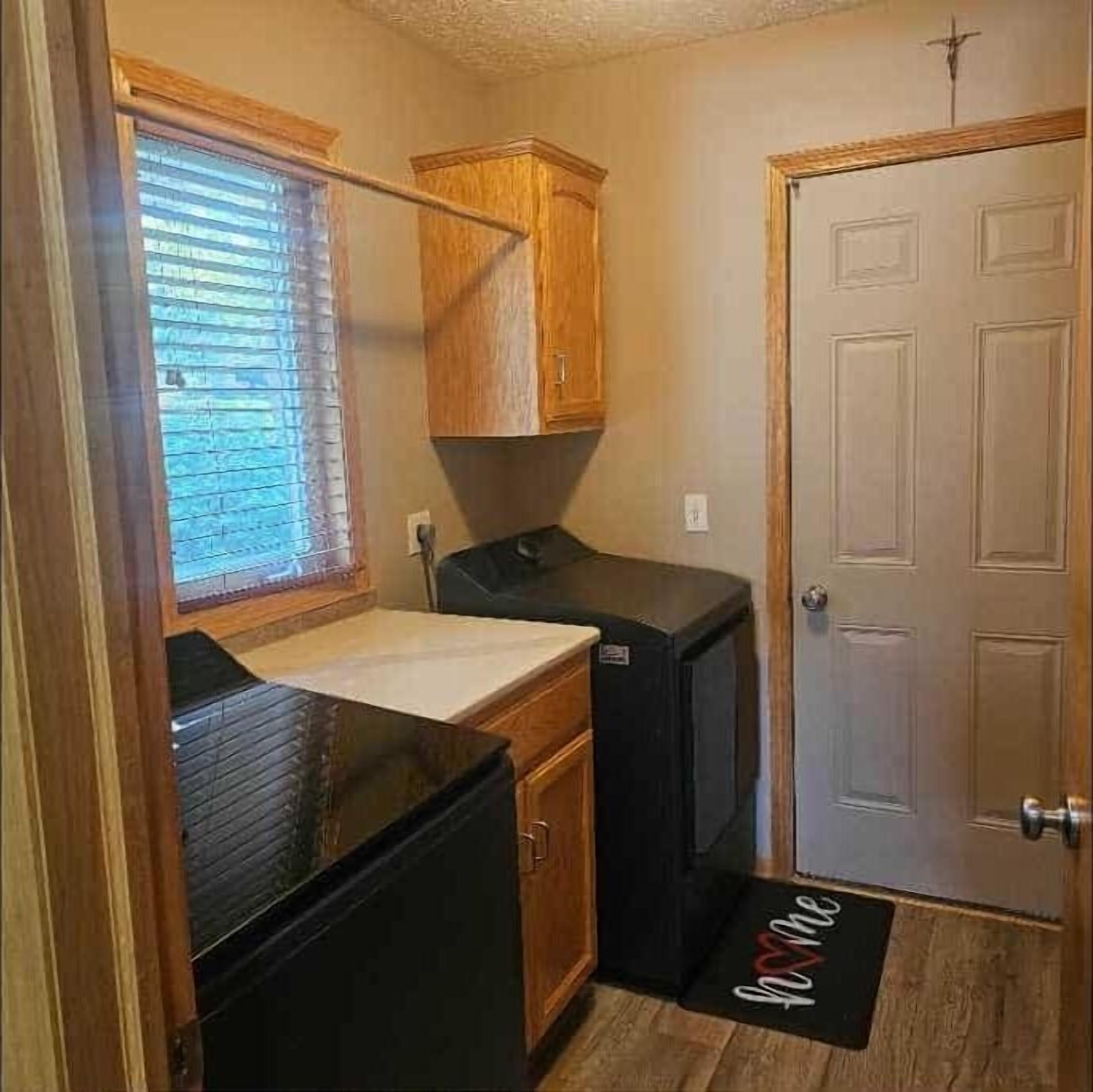 Une cuisine de taille réduite, avec des meubles de rangement en bois, un réfrigérateur noir et une fenêtre équipée de stores.