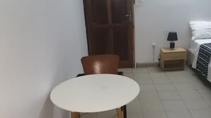 Et lille rum med et rundt bord og to stole, en trædør, et sengebord med en lampe og en seng med et mønstret dynedække.