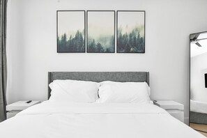 A modern bedroom with a large bed, white bedding, a grey headboard, and three framed forest landscape paintings on the wall.