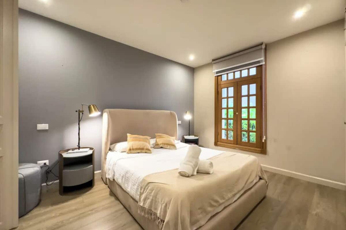 A bedroom with a large bed, bedside lamps, a nightstand, a window with a view, and a door.