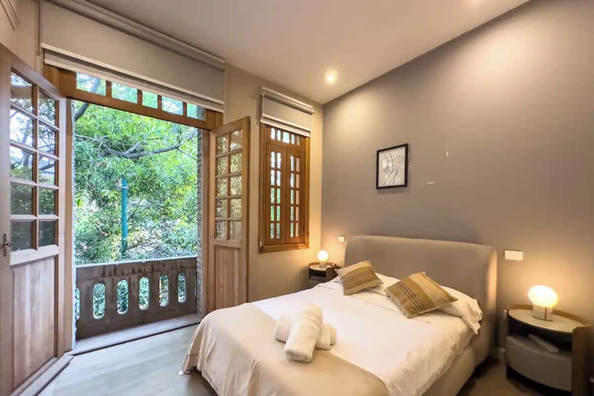 A bedroom with a large bed, a nightstand, a wall-mounted light, and a window with a view of greenery.