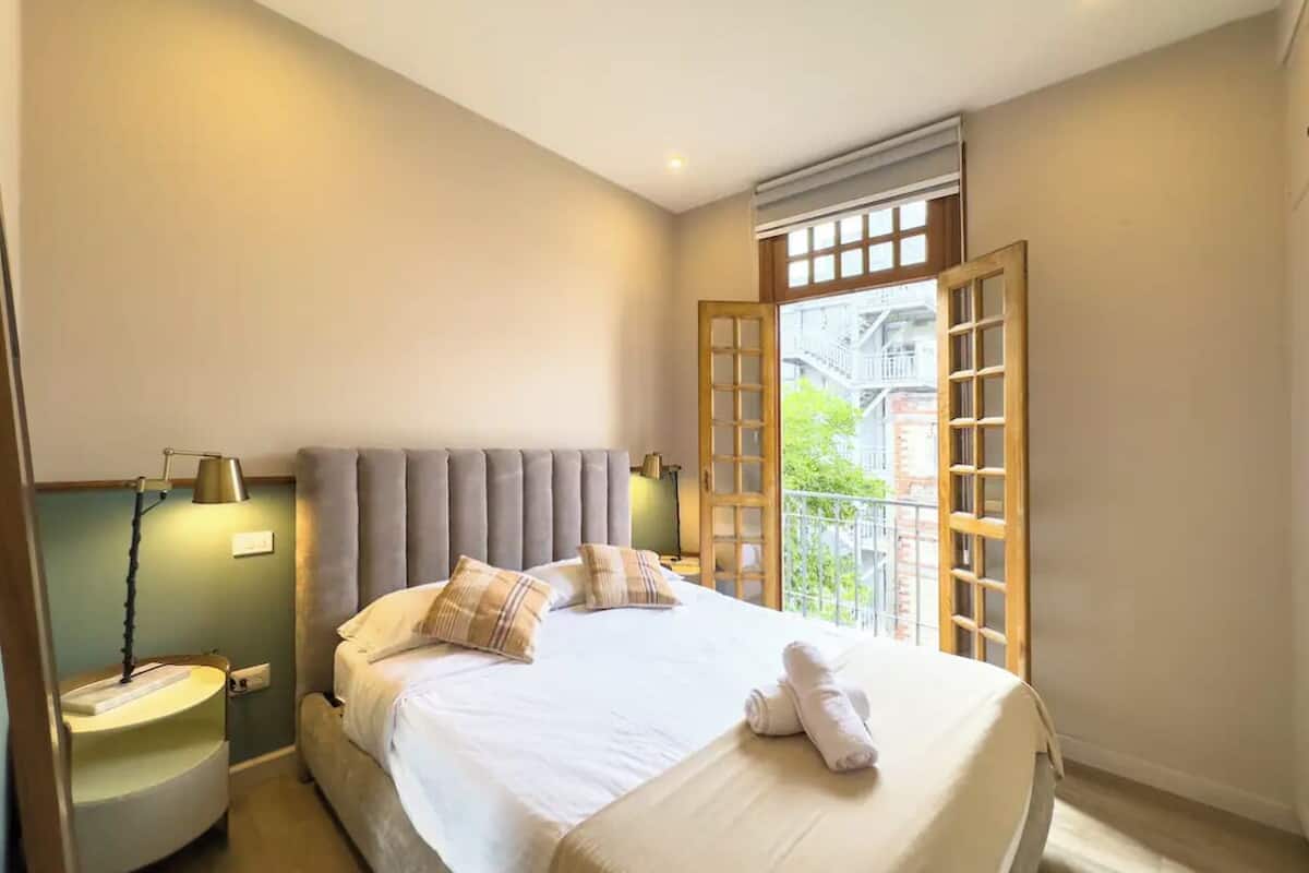 A neatly made bed with a headboard, a bedside lamp, and a window with a view of buildings.