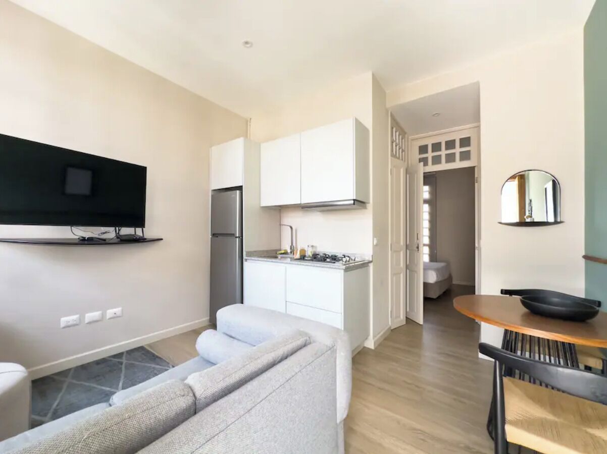 A modern living room with a grey sofa, a flat-screen TV mounted on the wall, and a wooden dining table with a bowl.