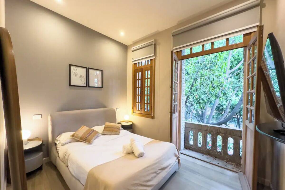 A bedroom with a large bed, bedside tables, a TV, and a balcony with a view of trees.