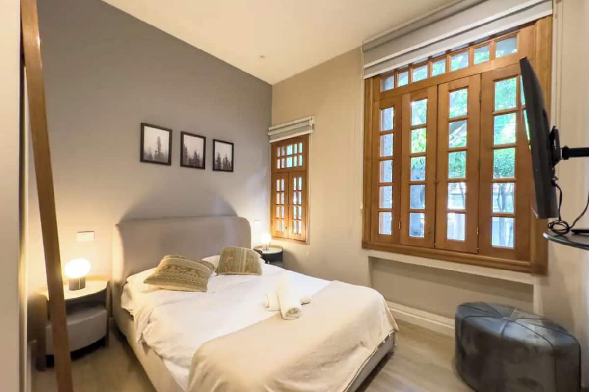 A bedroom with a bed, bedside tables, a window with blinds, and a television mounted on the wall.