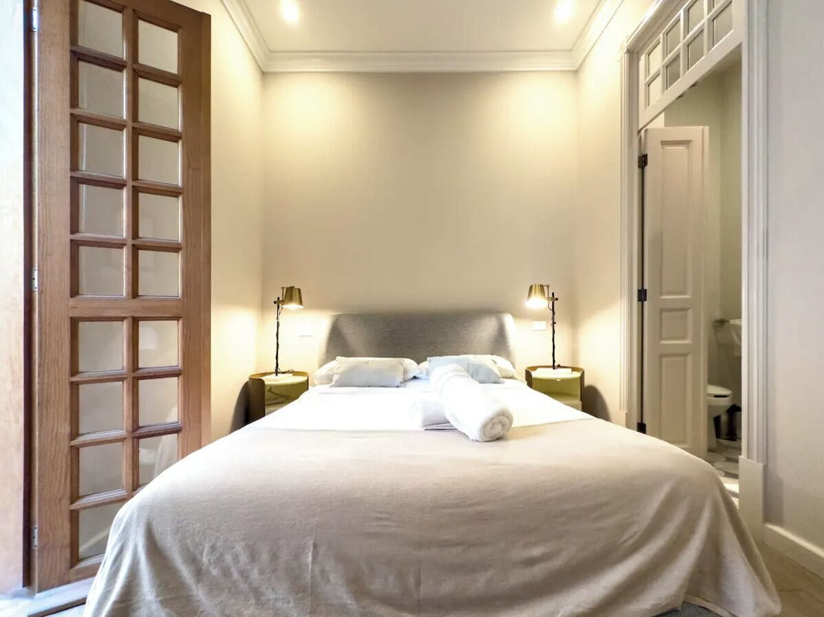 Una cama bien hecha con ropa blanca, dos mesitas de noche con lámparas y un espejo grande en la pared.