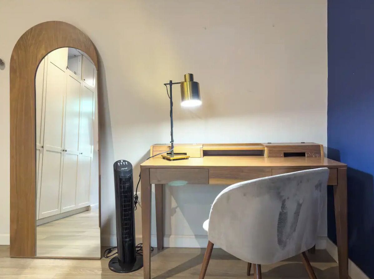 Una sala de estudio con escritorio, silla, lámpara y ventilador de pie.