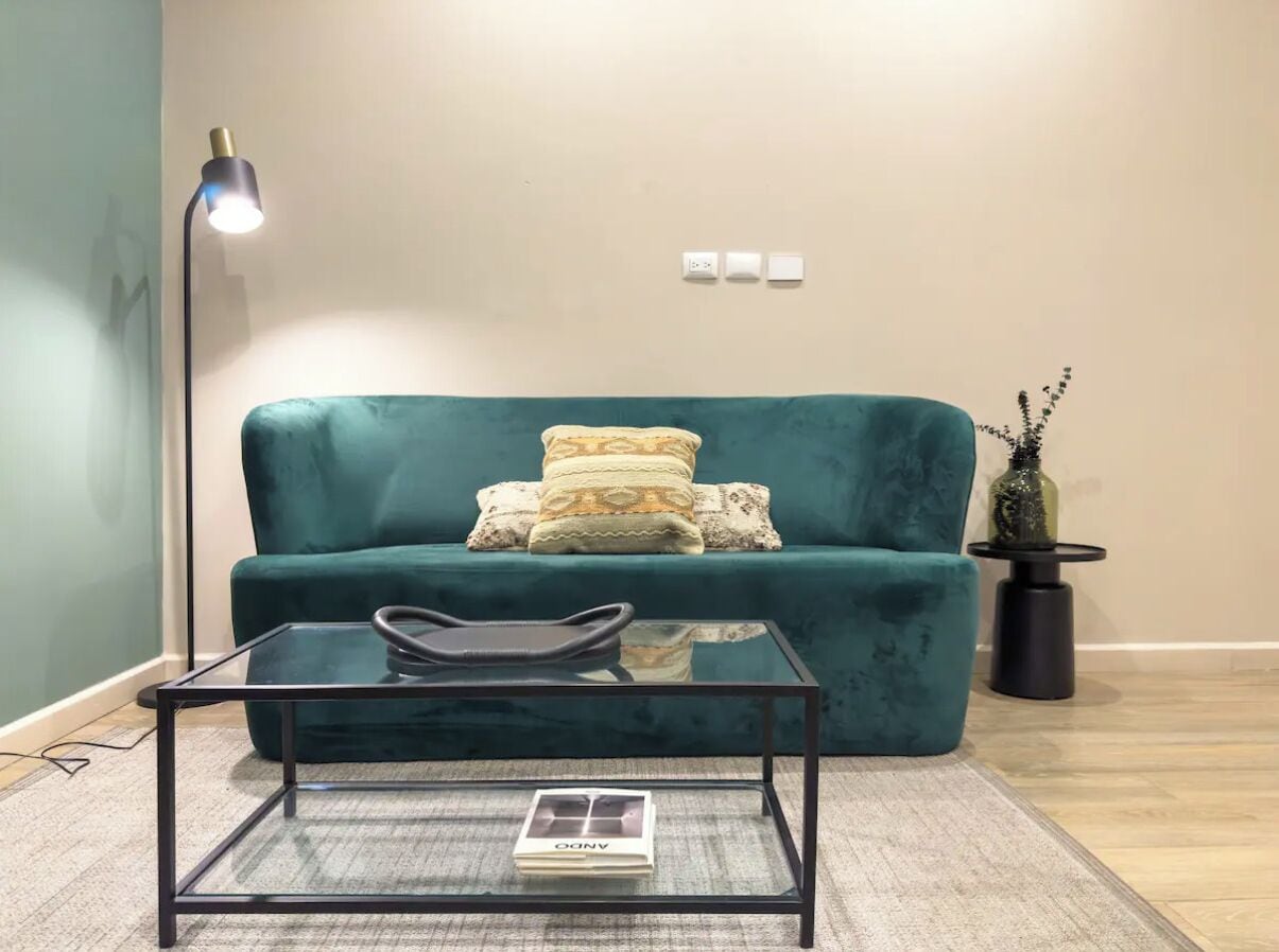 Un sofá color turquesa con cojines estampados, una mesa de centro de vidrio, una lámpara de pie y una pequeña planta en maceta.