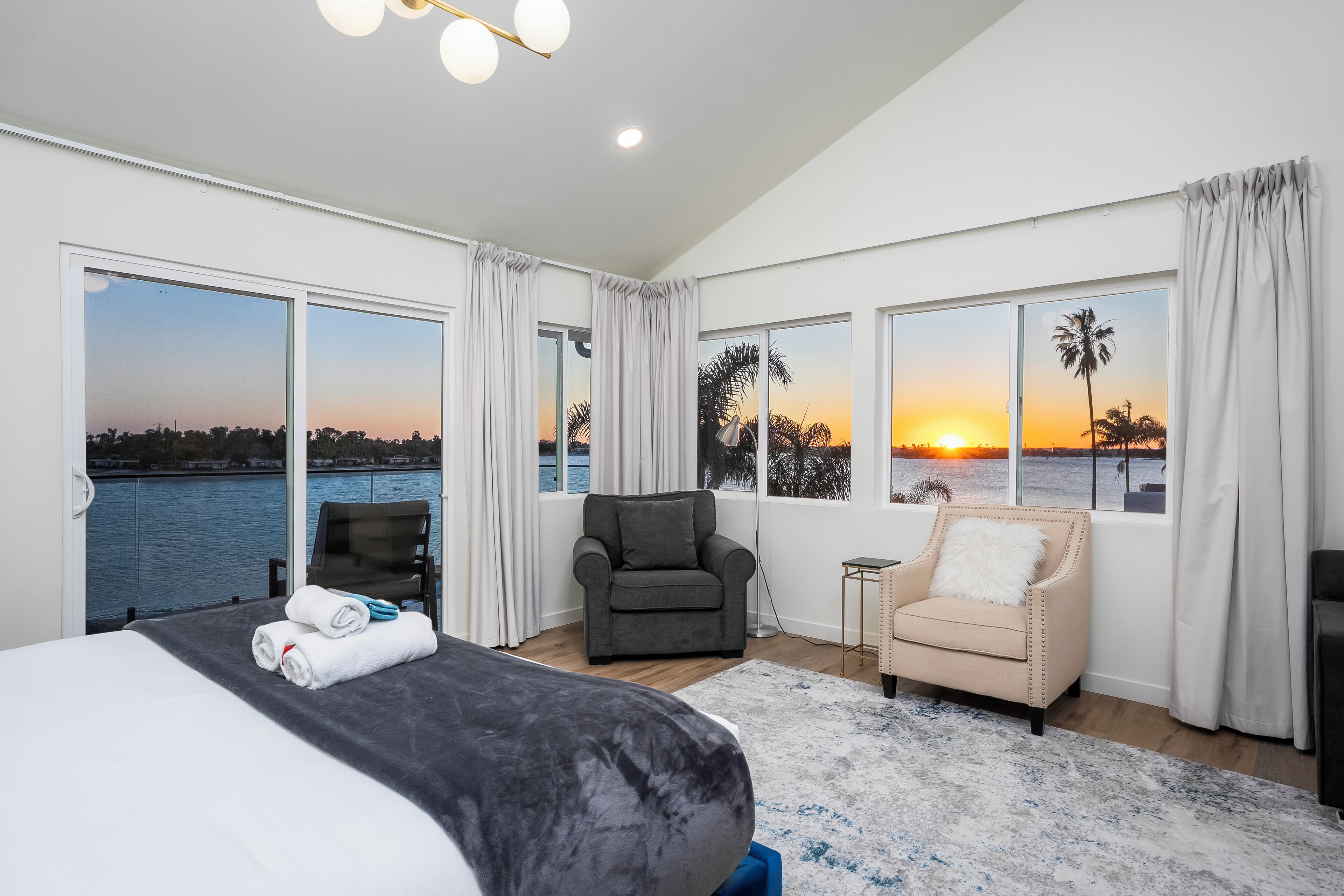 A spacious bedroom with a large bed, a grey armchair, a small side table, and a view of the sunset over water.