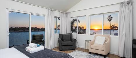 A spacious bedroom with a large bed, a grey armchair, a small side table, and a view of the sunset over water.
