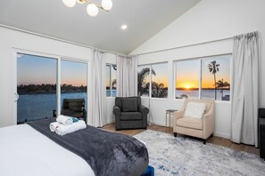 Quarto espaçoso com uma cama grande, uma poltrona cinza, uma mesinha de cabeceira e vista para o pôr do sol sobre a água.