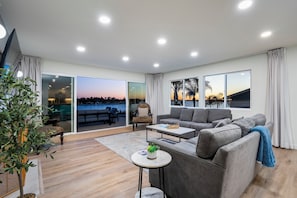 Uma sala de estar moderna com um sofá cinzento, uma mesa redonda pequena e uma grande janela com vista para o pôr do sol.