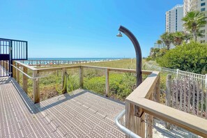 Eine Holzterrasse mit Metallgeländer, die auf einen Strand mit einer Palme und einem Gebäude im Hintergrund blickt.