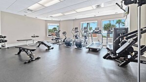 Un gimnasio con diverso equipo de ejercicio, ventanas amplias y vistas a palmeras.