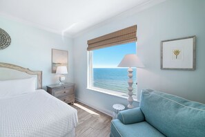 Ein Schlafzimmer mit einem Bett, einem Nachttisch, einer Lampe, einem Fenster mit Meerblick und einem gerahmten Bild an der Wand.