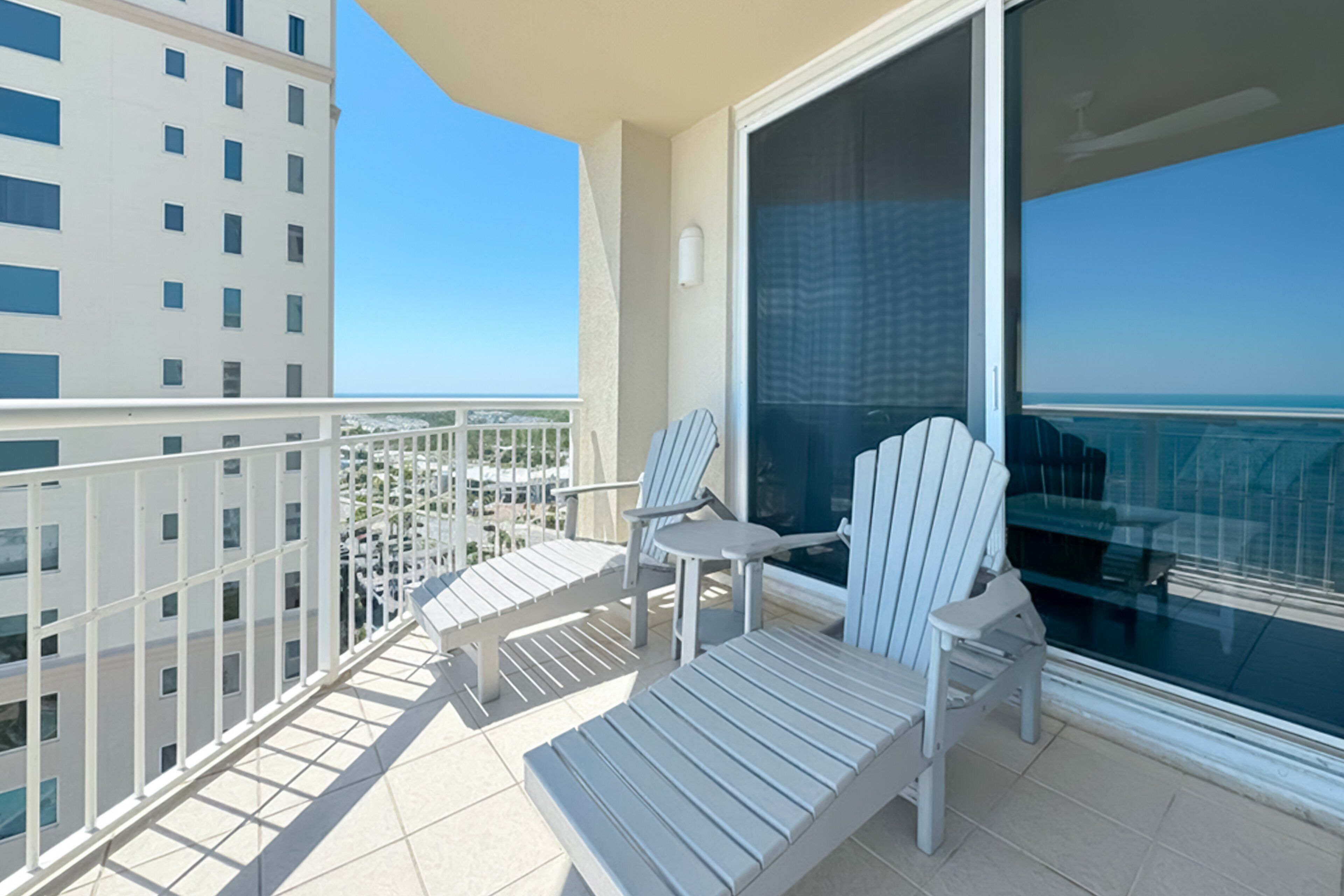 A balcony with two Adirondack chairs and a small table, overlooking a cityscape and a clear sky.