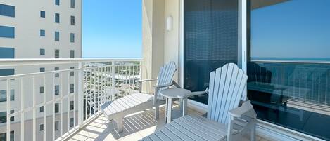 A balcony with two Adirondack chairs and a small table, overlooking a cityscape and a clear sky.