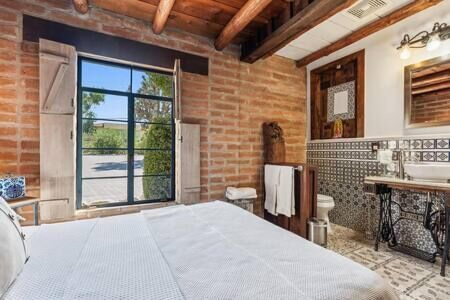 Une chambre dotée d’un grand lit, d’une porte-fenêtre coulissante donnant sur un balcon, d’une salle de bain avec un mur recouvert de carreaux et de poutres en bois au plafond.