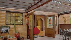 An open wooden door leading to a room with a red chair and a table. There are potted plants and a cactus outside.