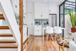 A modern kitchen with white cabinets, a stainless steel refrigerator, and a dining area with white chairs. - The Newtown Nest (Newtown)