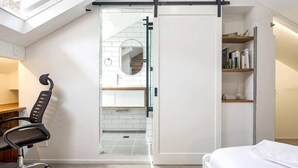 Un dormitorio moderno con una cama, un escritorio con una silla, una puerta corrediza a un baño y una estantería.