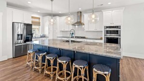 A modern kitchen with white cabinets, a stainless steel refrigerator, a large island with bar stools, and pendant lighting. - Isle Escape by Brunswick Vacation Rentals: Pool, Elevator & Stunning Ocean Views (Ocean Isle Beach)