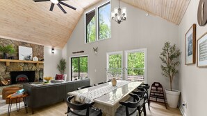A spacious living room with a high ceiling, a fireplace, a large dining table with chairs, and large windows offering a view of greenery.