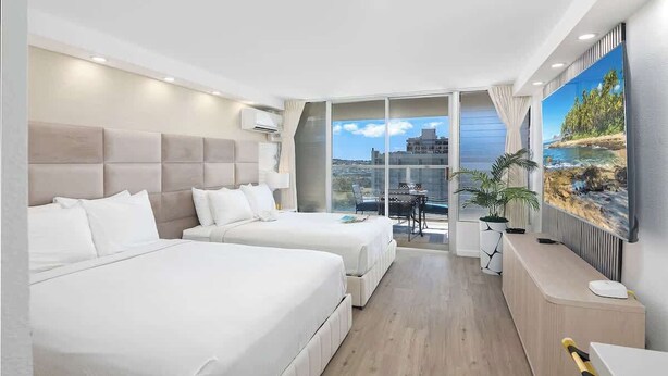 Een hotelkamer met twee bedden, een bureau, een televisie en een balkon met uitzicht op de stad.