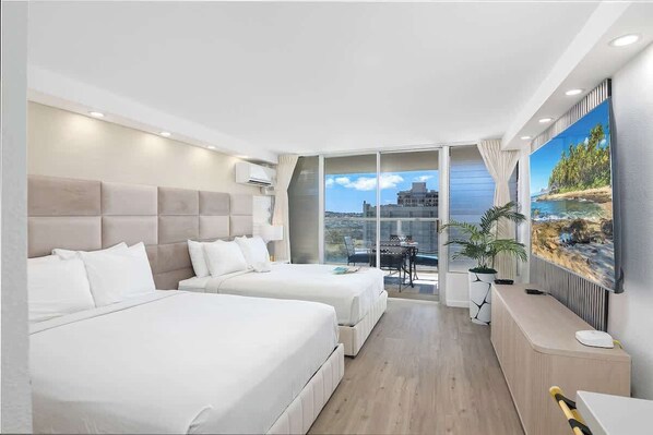 A hotel room with two beds, a desk, a TV, and a balcony with a city view.