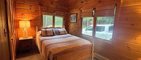 A wooden cabin bedroom with a bed, a nightstand, a lamp, and windows overlooking a forest.